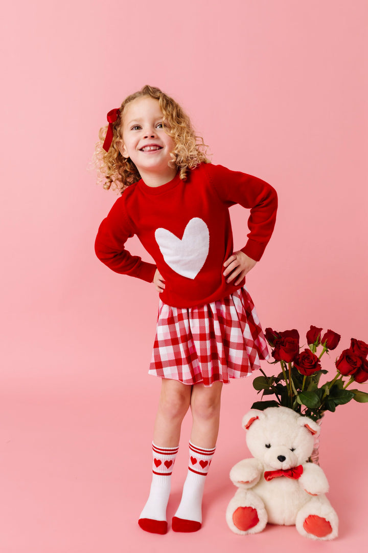 young girl standing by stuffed bear and red roses wearing bright red heart sweater paired with red and pink plaid skort and valentines themed crew socks