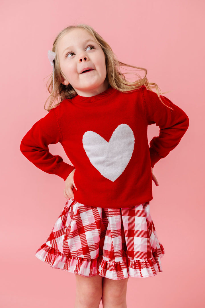 little girl in red cotton knit pullover sweater with white heart design layered over pink, red and white plaid twirl dress for Valentine's day