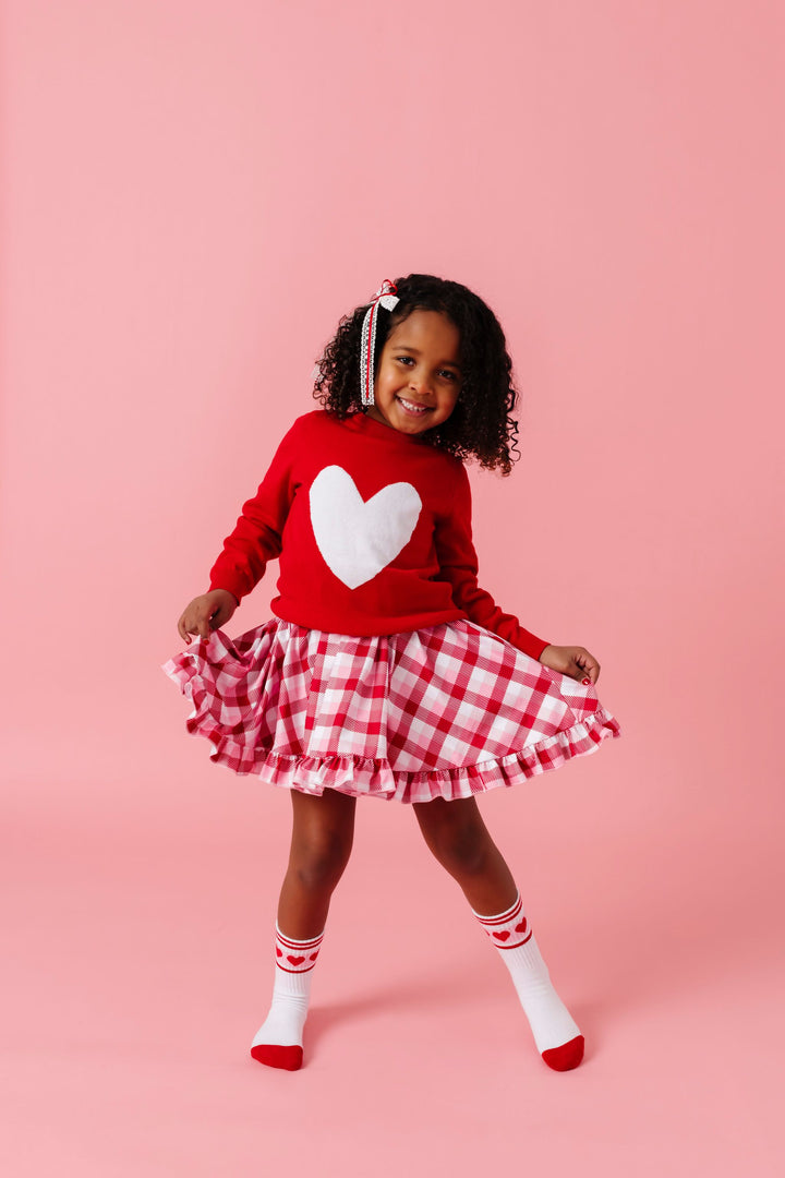 young girl in valentines plaid twirl dress paired with red sweater with white heart design, matching heart crew socks and white lace with red ribbon hair bow
