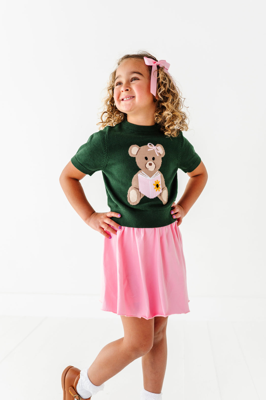 young girl in forest green short sleeve sweater with bear holding a book design and matching light pink twirl skort and hair bow