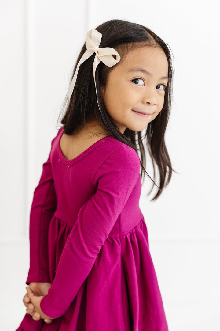 little girl looking over shoulder to show scoop back for magenta long sleeve twirl dress and cream satin hair bow on clip