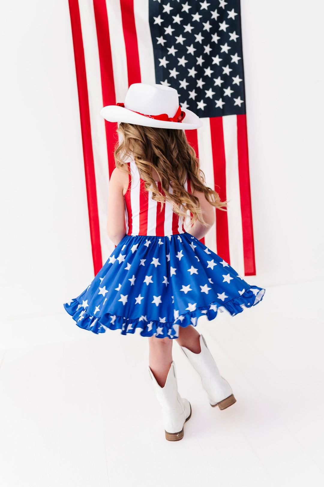 little girl twirling in front of American flag in red, white and blue flag inspired twirl dress paired with cowgirl hat and boots