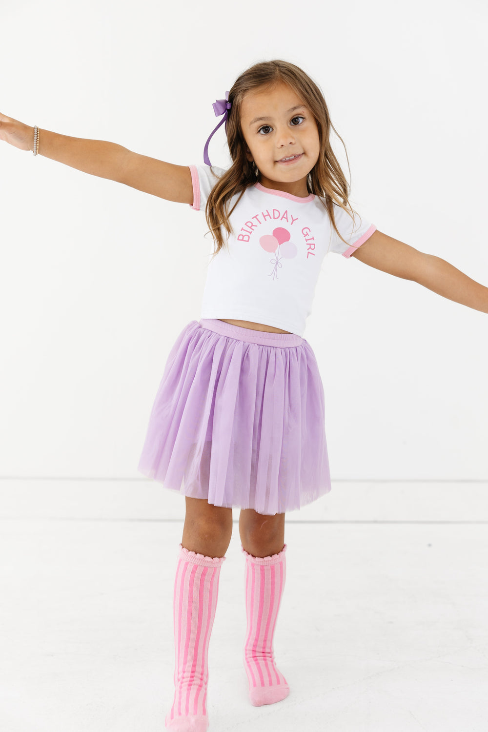 little girl in white fitted tee with pink binding and Birthday Girl balloon print paired with lavender tulle skort, bow and pink striped knee socks