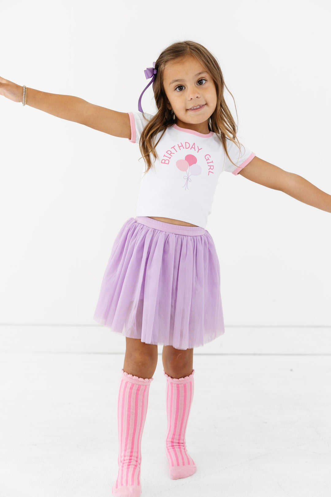 little girl in white fitted tee with pink binding and Birthday Girl balloon print paired with lavender tulle skort, bow and pink striped knee socks