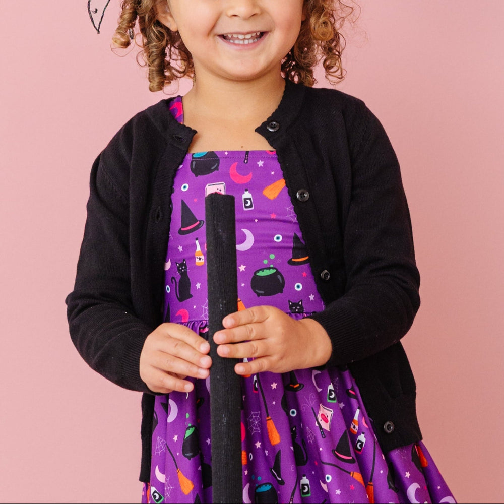 little girl holding broom in black cotton knit cardigan with buttons and purple twirl dress with witchy themed print