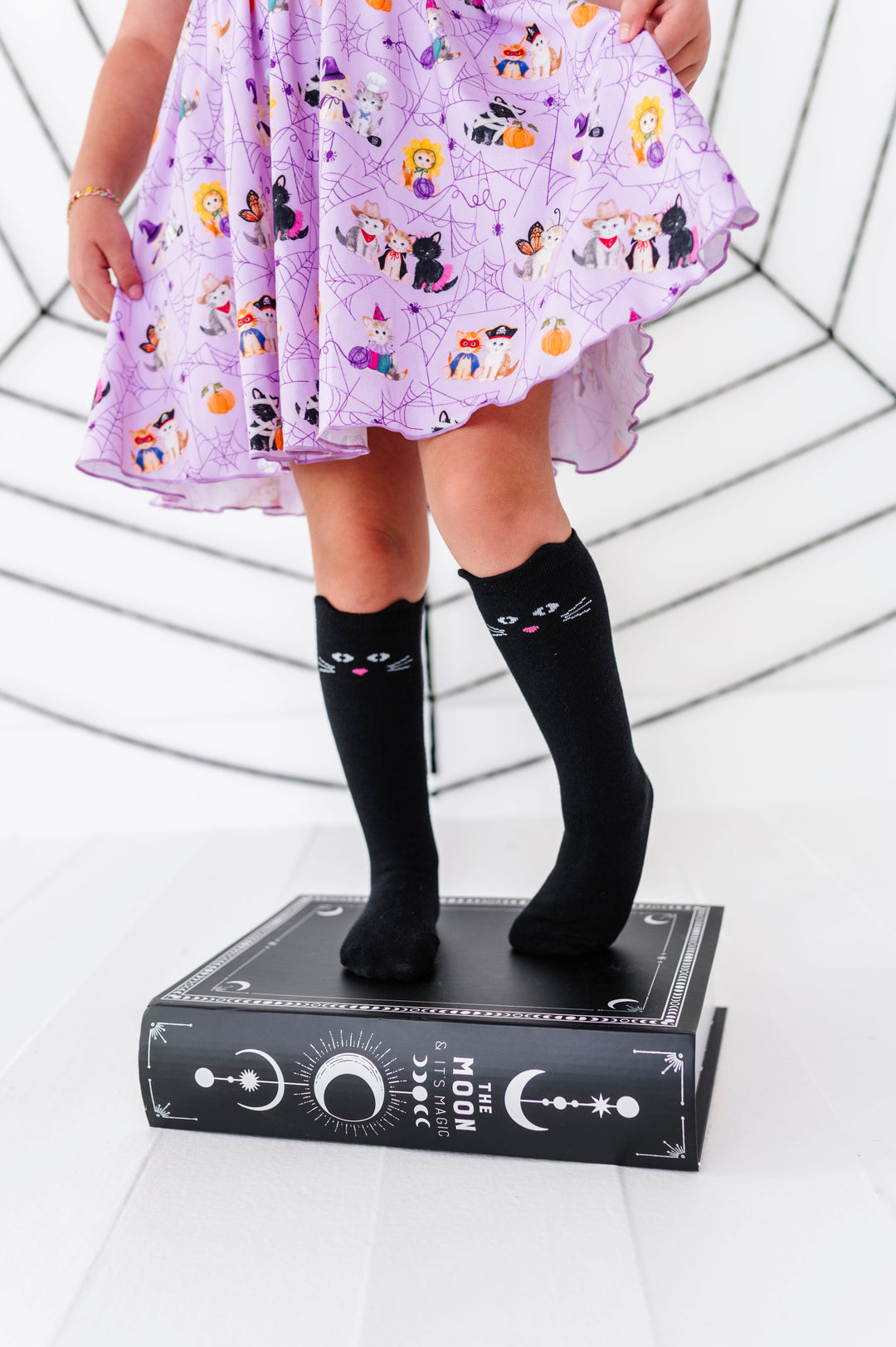 little girl standing on spell book in lavender dress printed with kittens wearing Halloween costumes and paired with black knee socks with cat ears and face design on the front