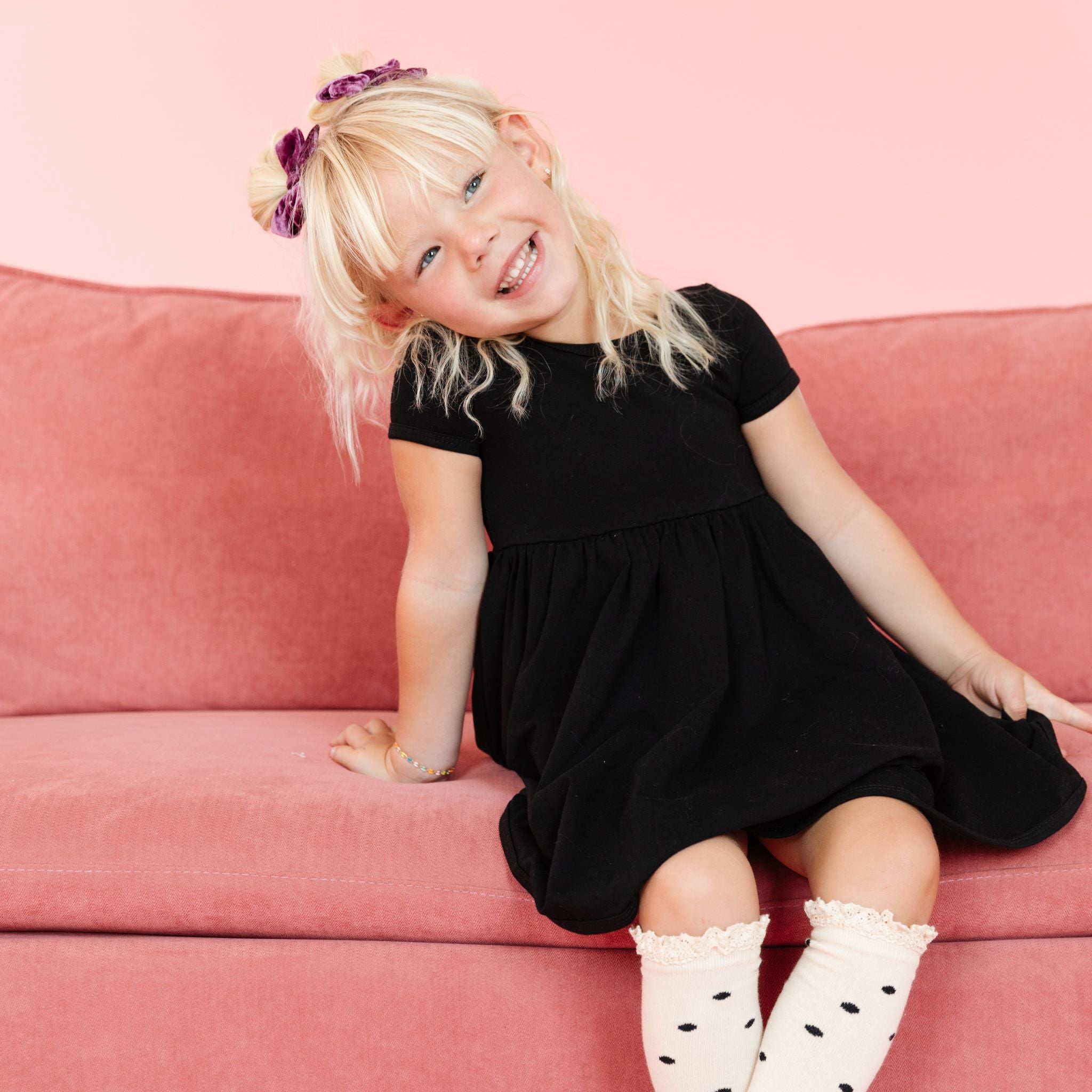 little girl in classic black fall dress with matching cream and black dot knee high socjs and purple velvet pigtail bows