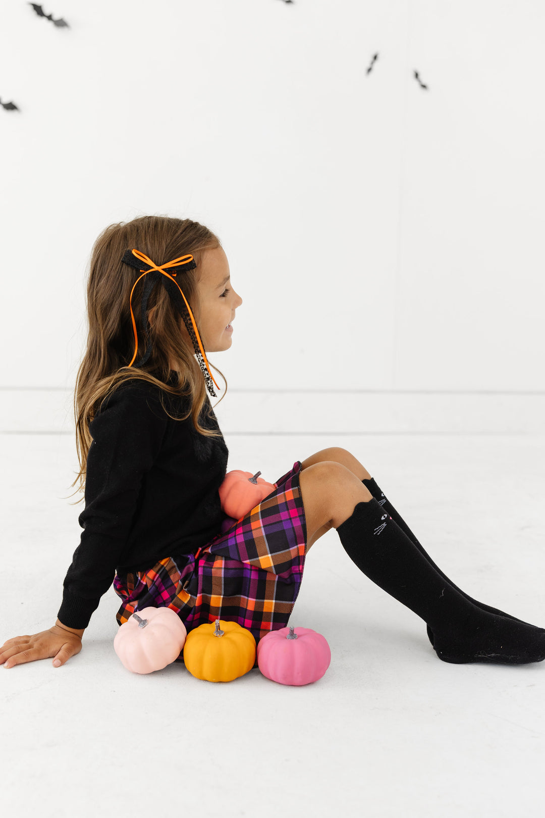 little girl sitting with colorful pumpkins in October plaid skort and black sweater with matching black lace bow with orange ribbon trim