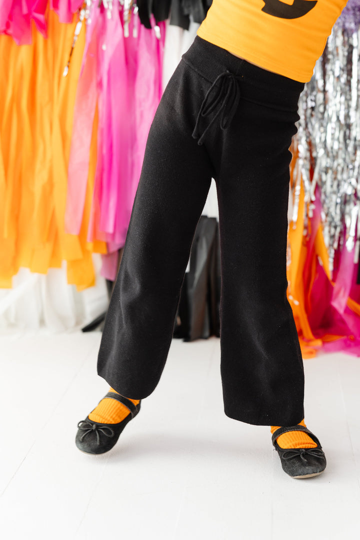 closeup of little girl in 100% cotton knit black sweater pants with drawstring waistband paired with orange top and ribbed midi socks with black mary janes