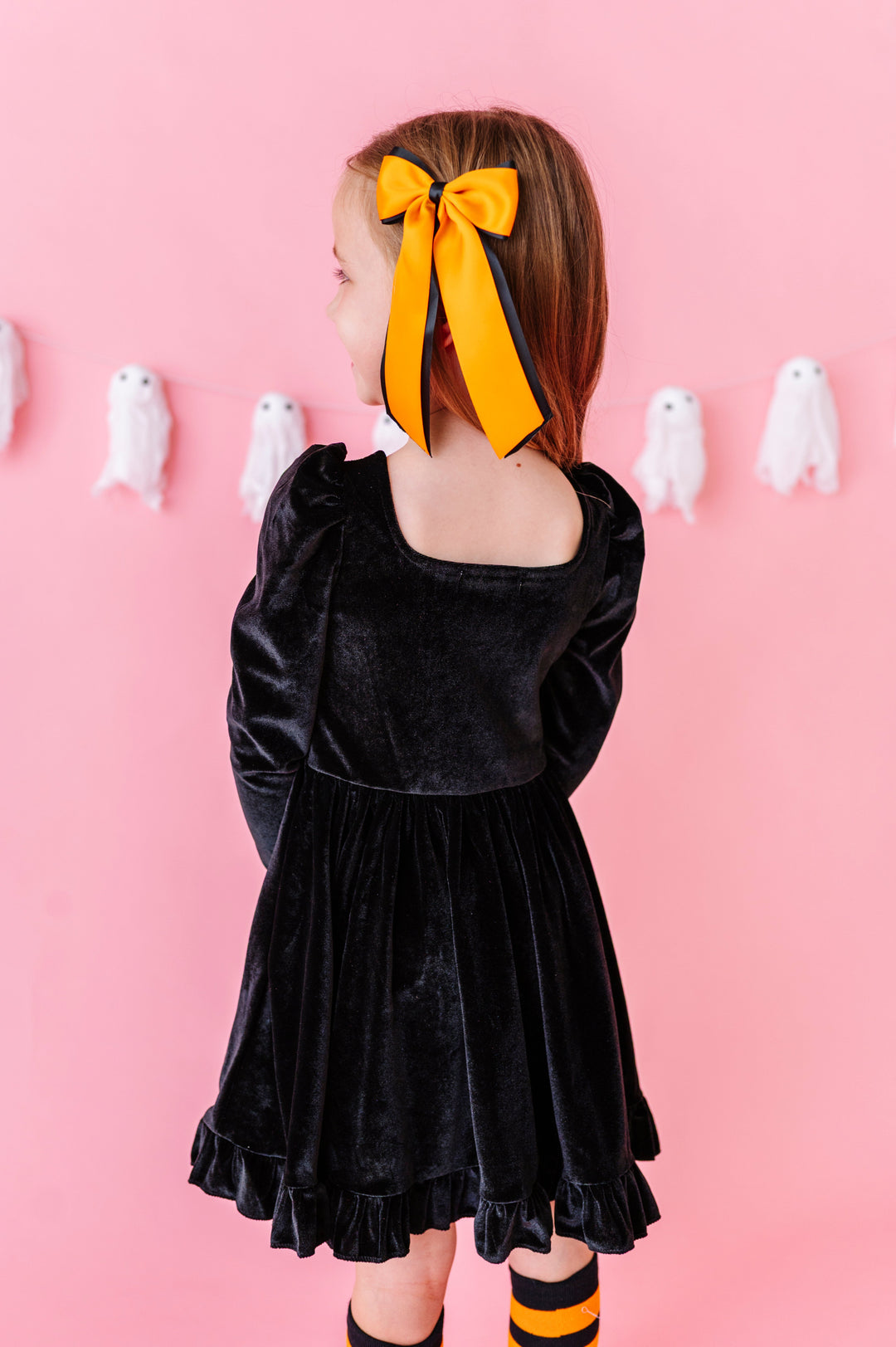 back view of little girl in front of pink background with ghost banner wearing black velvet long sleeve twirl dress with square neckline and ruffled trim paired with orange and black ribbon Halloween hair bow