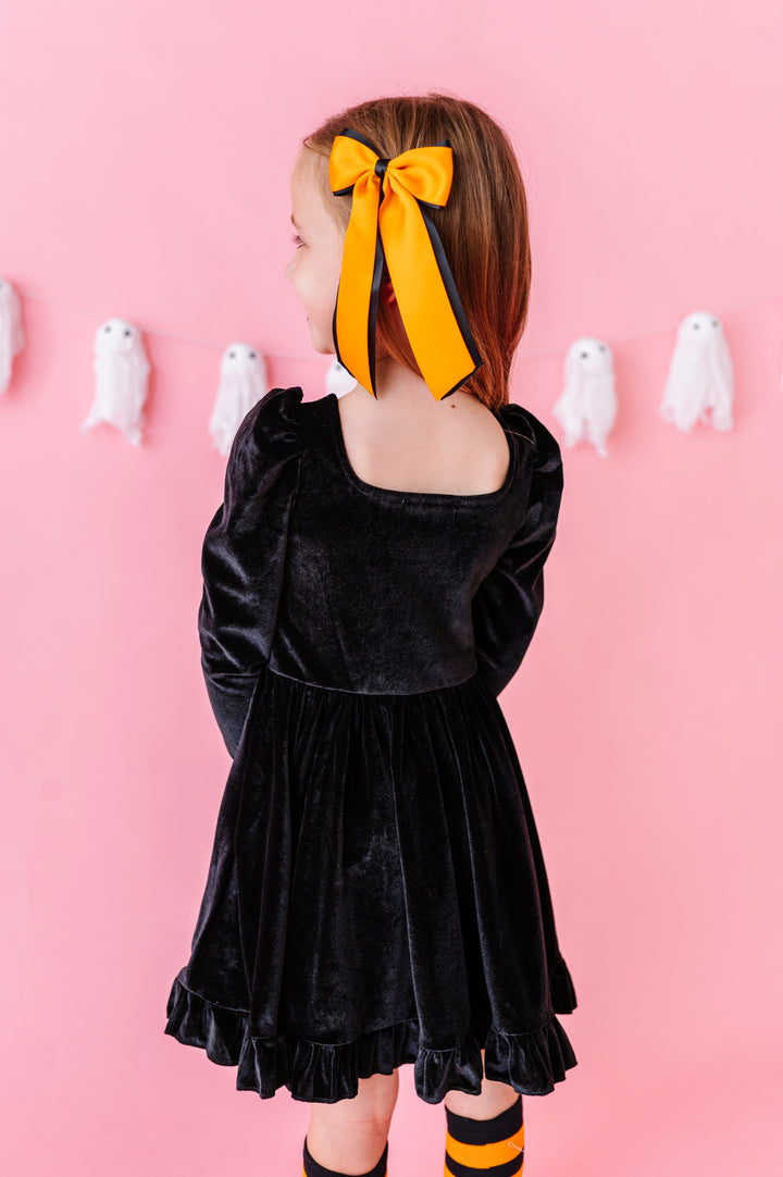 back view of little girl in front of pink background with ghost banner wearing black velvet long sleeve twirl dress with square neckline and ruffled trim paired with orange and black ribbon Halloween hair bow