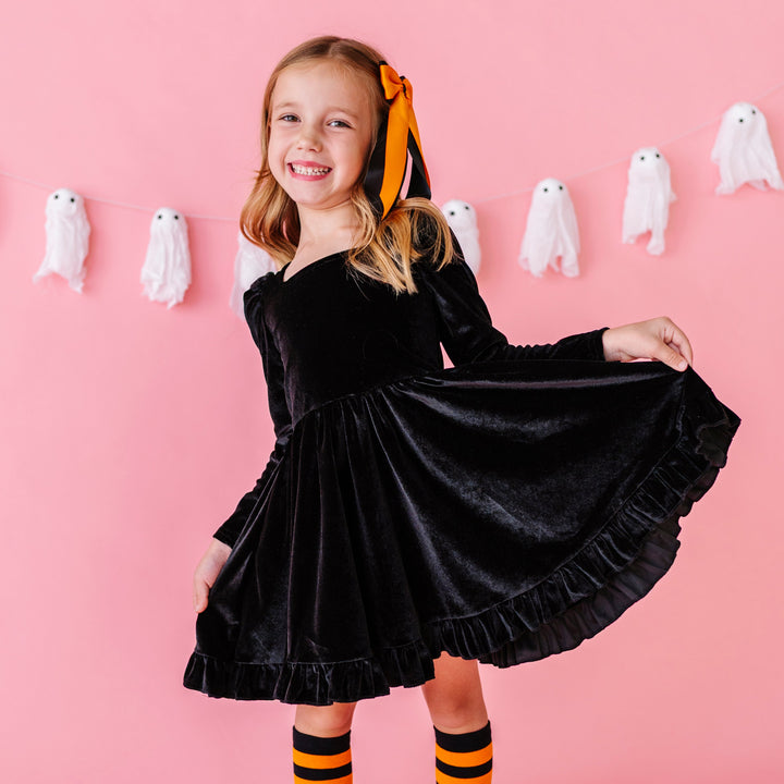 young girl in front of light pink background with ghost banner wearing black velvet long sleeve twirl dress paired with orange and black ribbon bow and matching striped knee socks