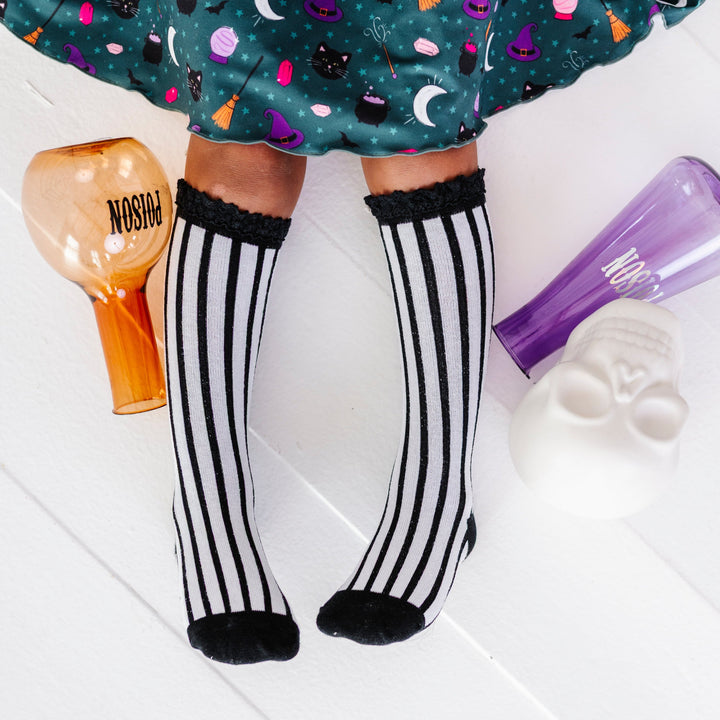 little girl sitting with potion bottles in black and white vertical striped knee socks with lace tops paired with wtitch print twirl dress