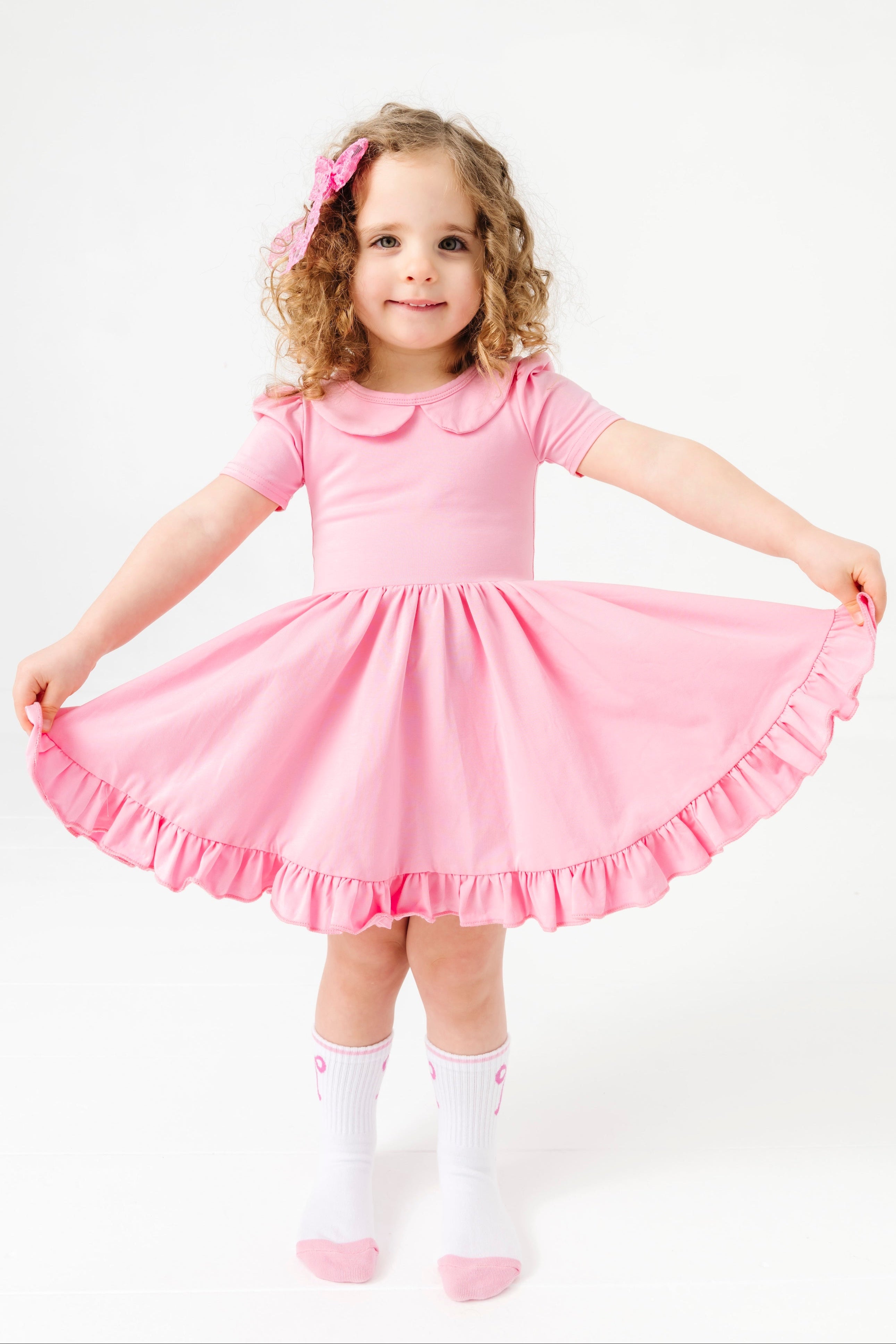 little girl in solid pastel pink short sleeve twirl dress with peter pan collar and matching pink bow and crew socks with pink bow design