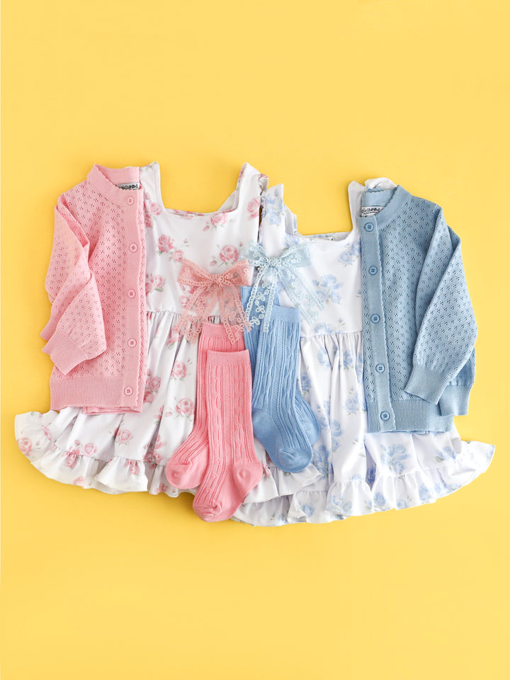 side by side flat lay of girl's white spring dresses with blossom and pastel blue floral prints paired with matching pointelle cardigans, cable knit knee high socks and lace hair bows