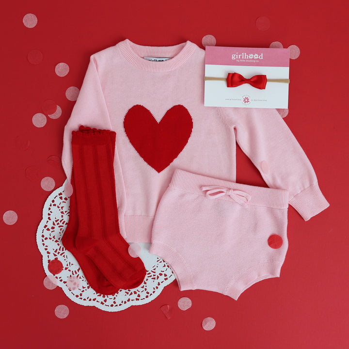 flatlay Valentine's Day sweater set outfit for baby girl in pink with red heart and matching bright red socks and bow