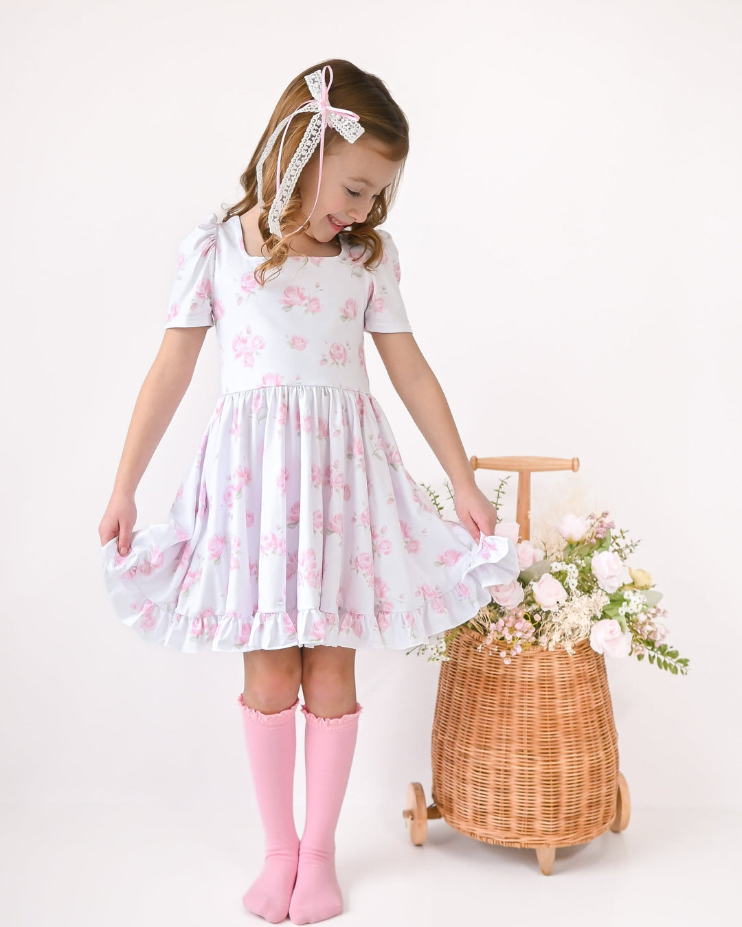 little girl with flower basket of roses wearing white twirl dress with pretty pink rose print paired with light pink socks and bow