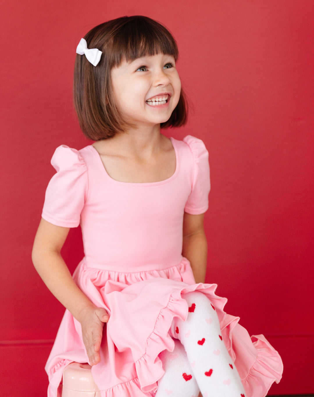 little girl sitting in square neck pastel pink twirl dress and matching red and pink heart print tights