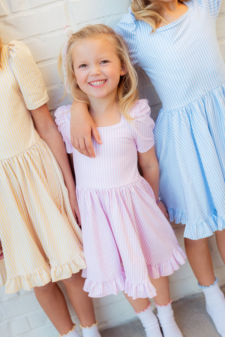 little girl in light pink and white pinstripe mod twirl dress and matching lettuce trim midi socks beside sisters in coordinating pinstripe dresses in yellow and blue