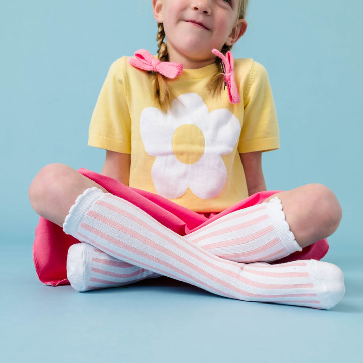 little girl sitting in pastel yellow knit sweater top with flower design paired with pink skort and pink and white scalloped knee socks