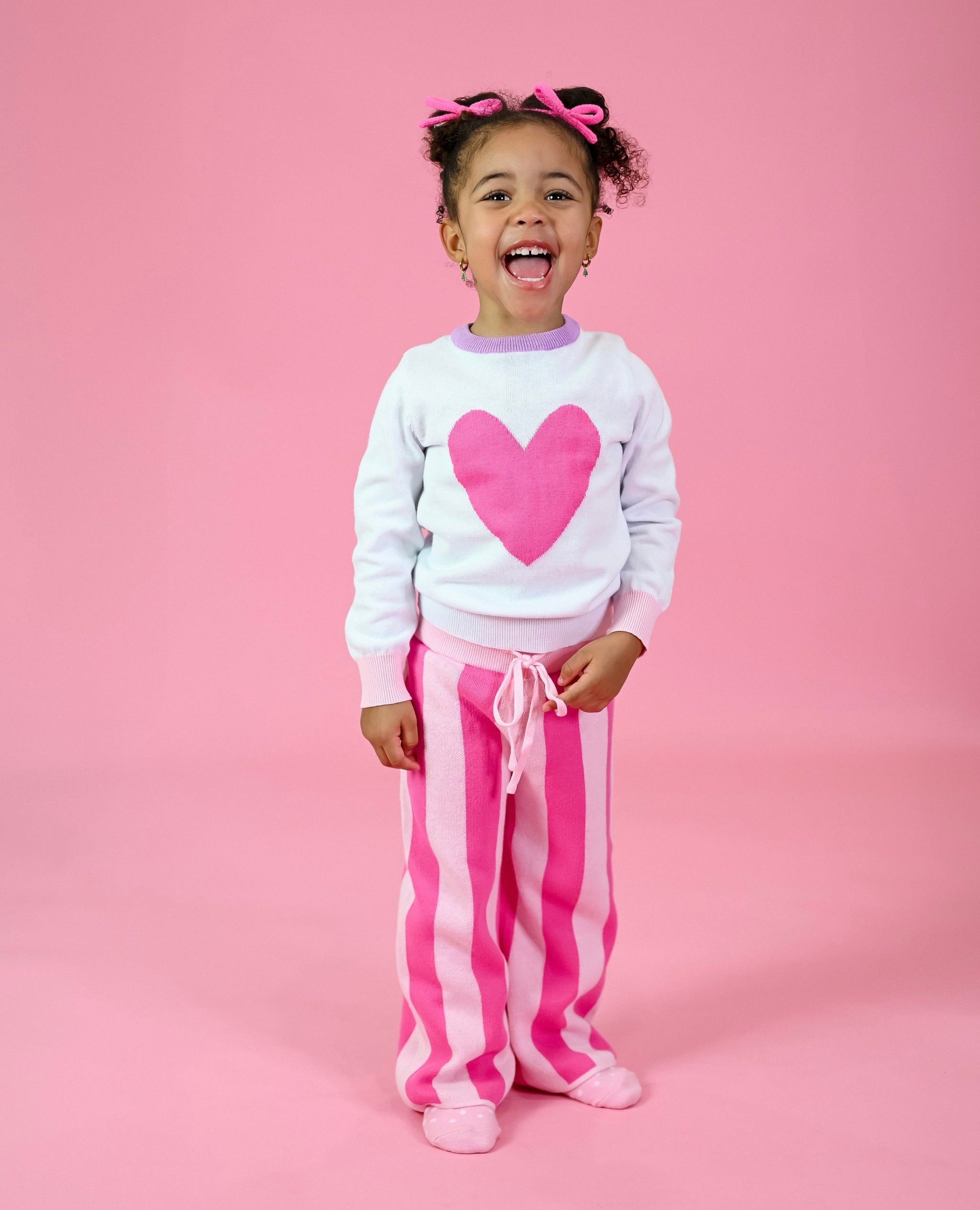 little girl in Valentine's themed sweater set featuring pullover sweater with pink heart and matching two tone pink striped pants and pink yarn pigtail bows