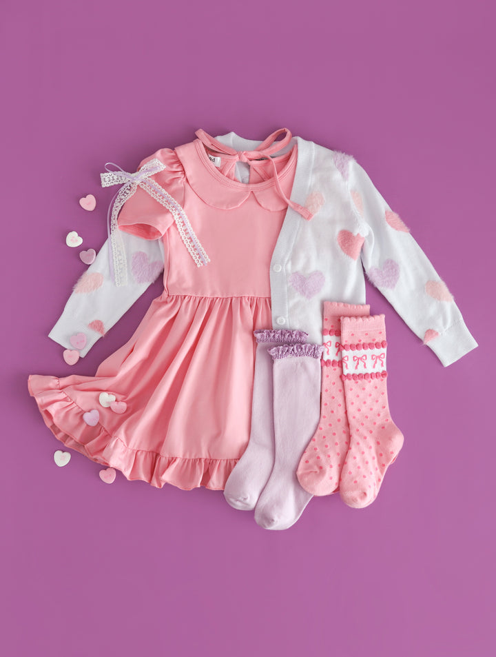 girls pastel Valentine's outfit with light pink twirl dress, white knit cardi with pastel fuzzy hearts and matching socks and bow doe conversation hearts vibes