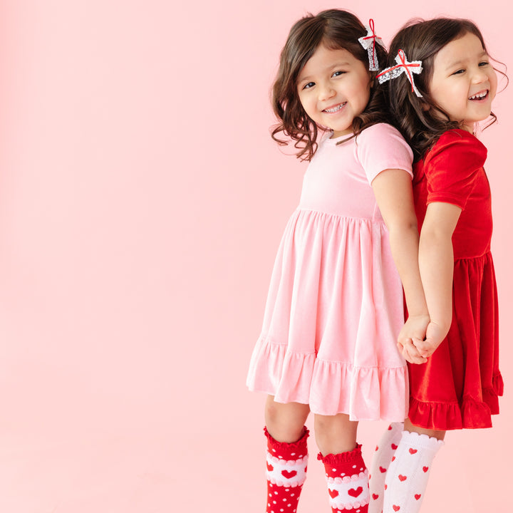 girls standing back to back in coordinating pink and red velvet twirl dresses styled with matching socks and bows for Valentine's Day