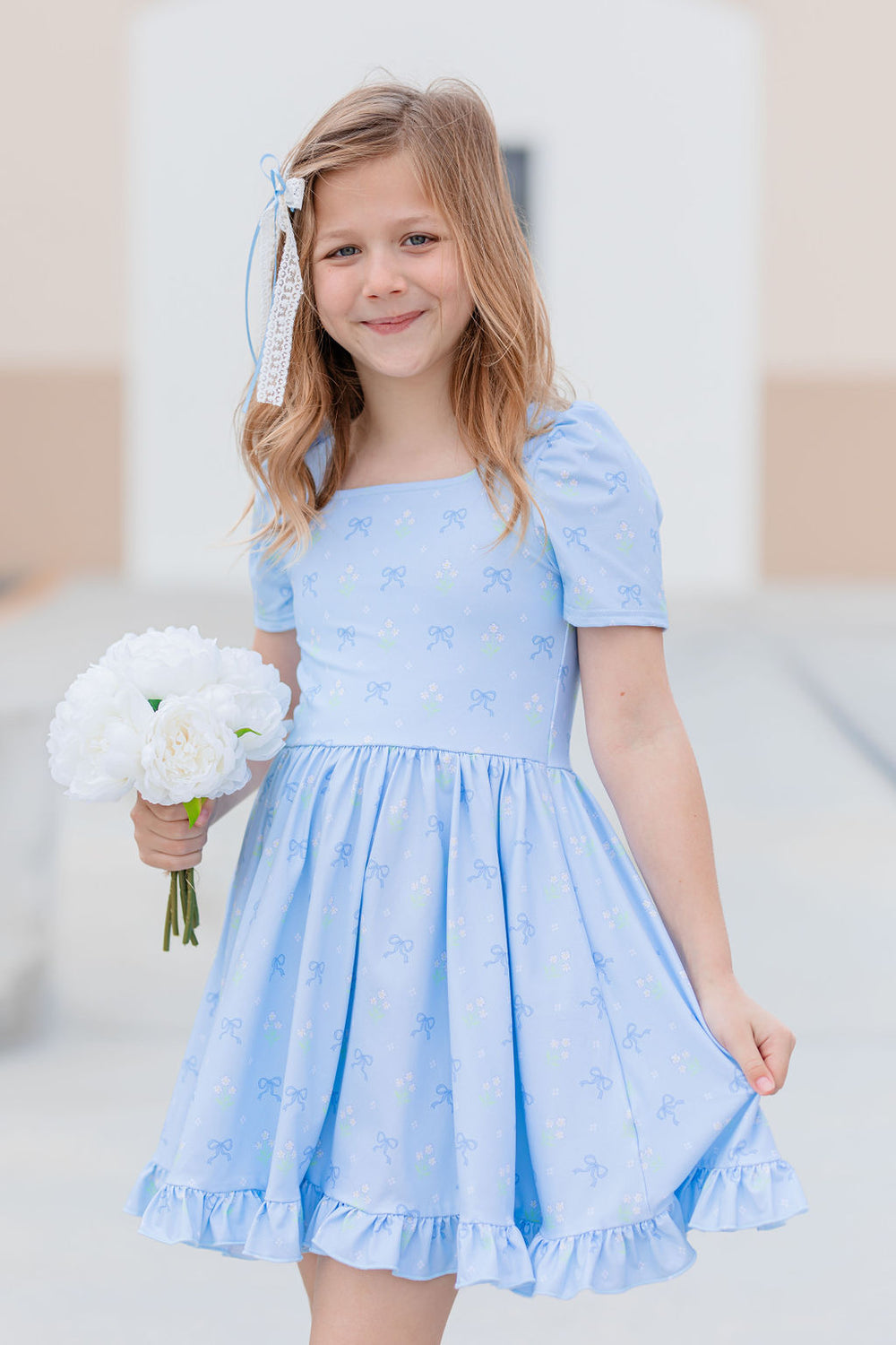 girl holding bouquet in blue square neck twirl dress with dainty bow and floral print and matching lace and ribbon hair bow 