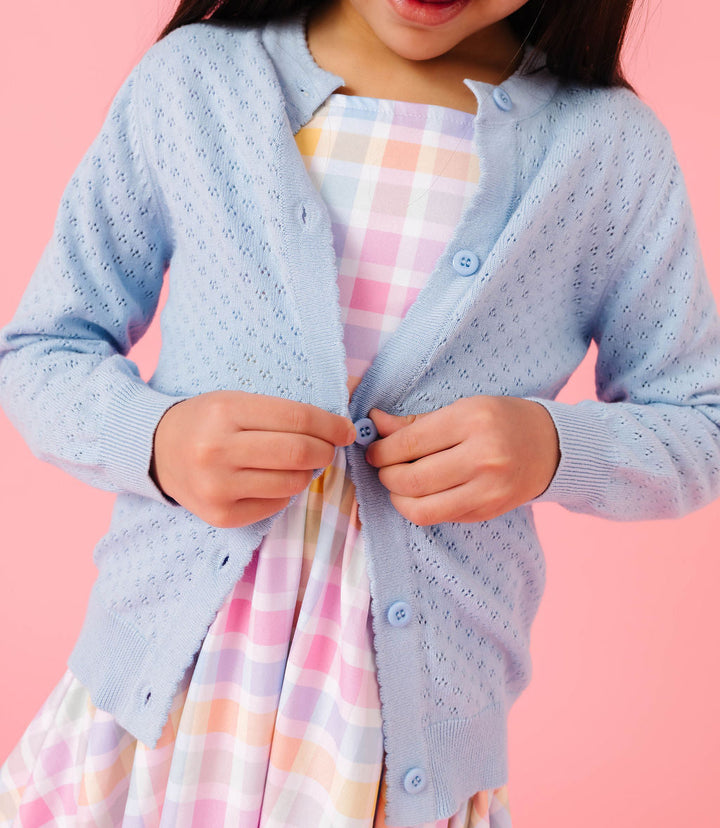 Child wearing a light blue cardigan over a pastel checkered dress on a pink background