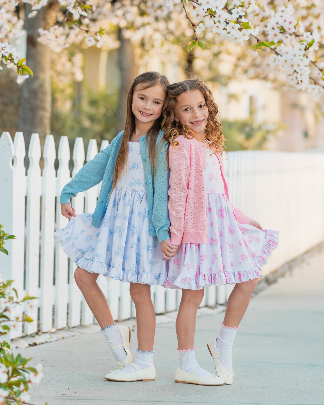 two girls in white spring floral twirl dresses, one with blue flowers and matching blue cardigan and lettuce trim midi socks and one with pink floral and matching pink pointelle cardigan and socks