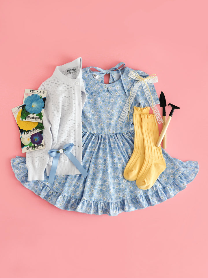 flat lay of girls' garden party outfit with blue micro floral collared twirl dress, white cotton knit pointelle cardigan, pastel yellow scallop trimmed knee socks and white lace bow with yellow satin ribbon