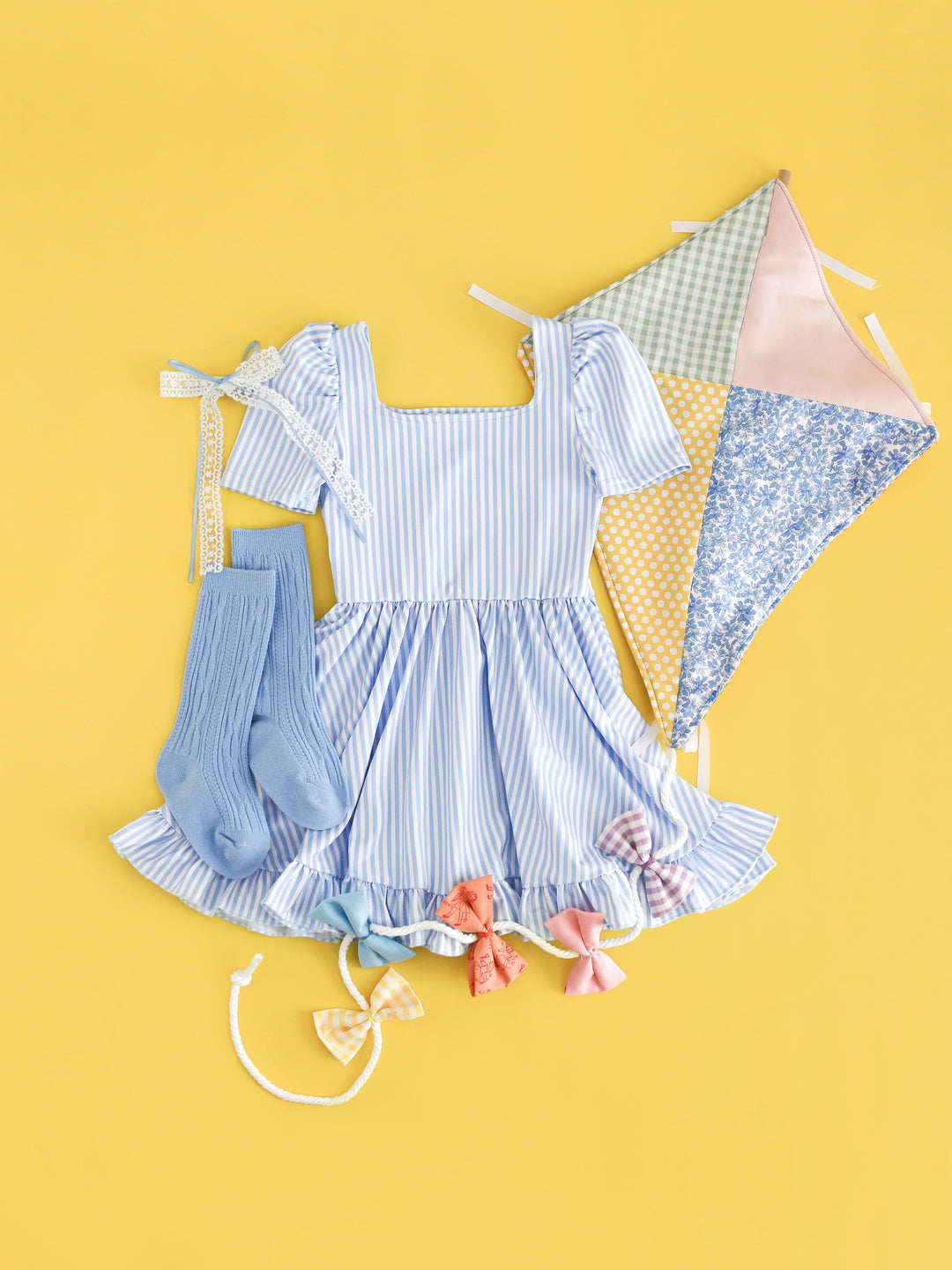 flat lay with patchwork kite and girls' light blue and white pinstripe square neck twirl dress with matching cable knit knee highs and bow