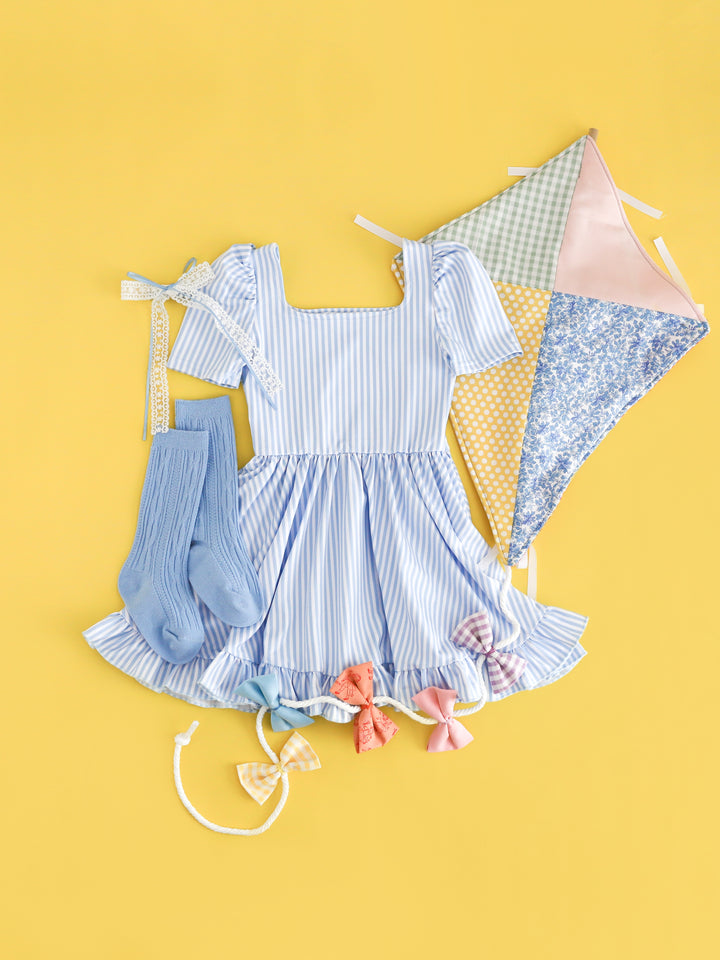 flat lay with patchwork kite and girls' light blue and white pinstripe square neck twirl dress with matching cable knit knee highs and bow