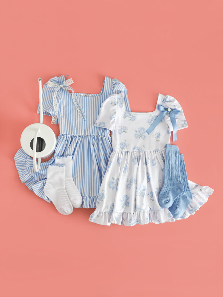 flat lay of coordinating white and blue spring twirl dresses for sister set; one with pinstripe print and one with dreamy rose print paired with matching socks and bows