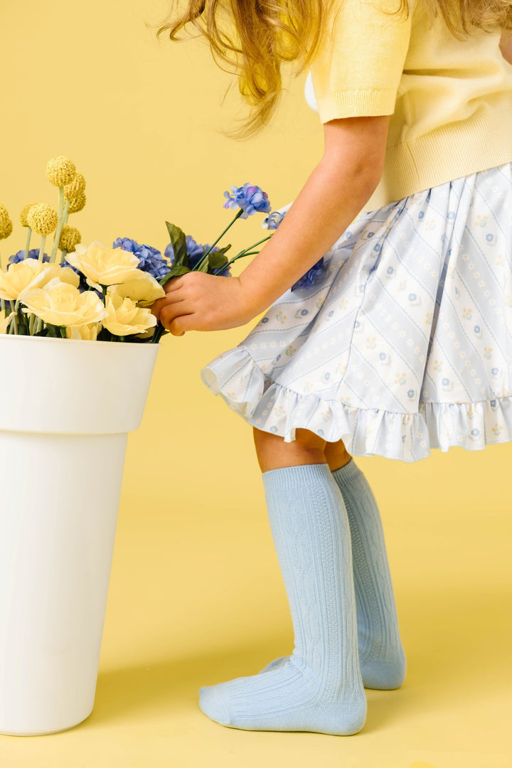 little girl picking flowers in pastel blue and yellow spring wallpaper print twirl dress paired with light blue cable knit knee highs and pastel yellow sweater top