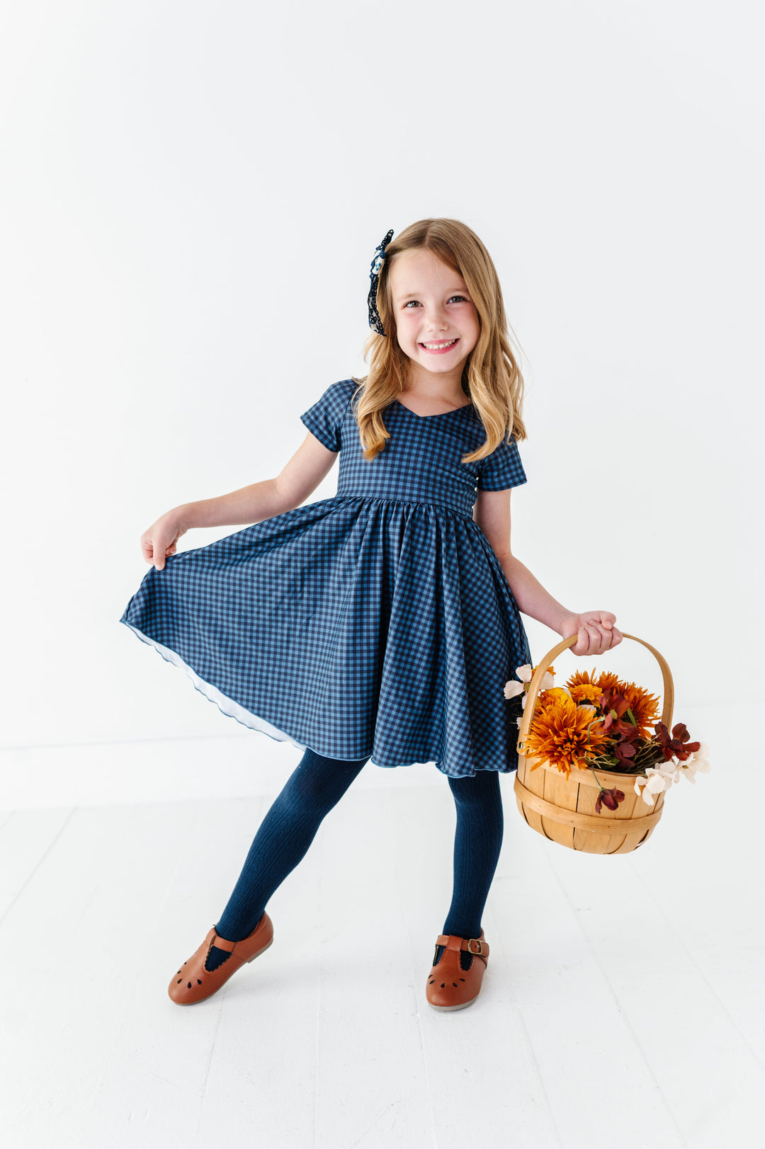 young girl holding fall flower basket in two-tone blue gingham v-neck twirl dress with lettuce trim paired with navy cable knit tights