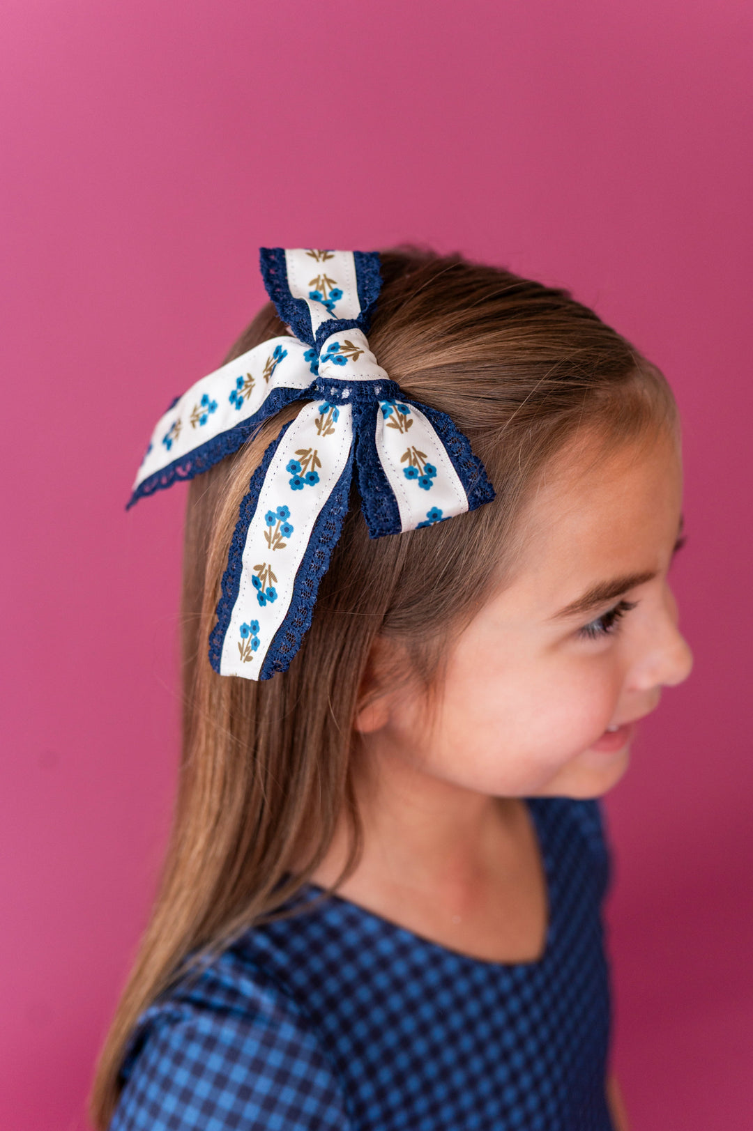 little girl photographed on pink background wearing blue floral hair bow with navy lace trim paired with blue gingham twirl dress