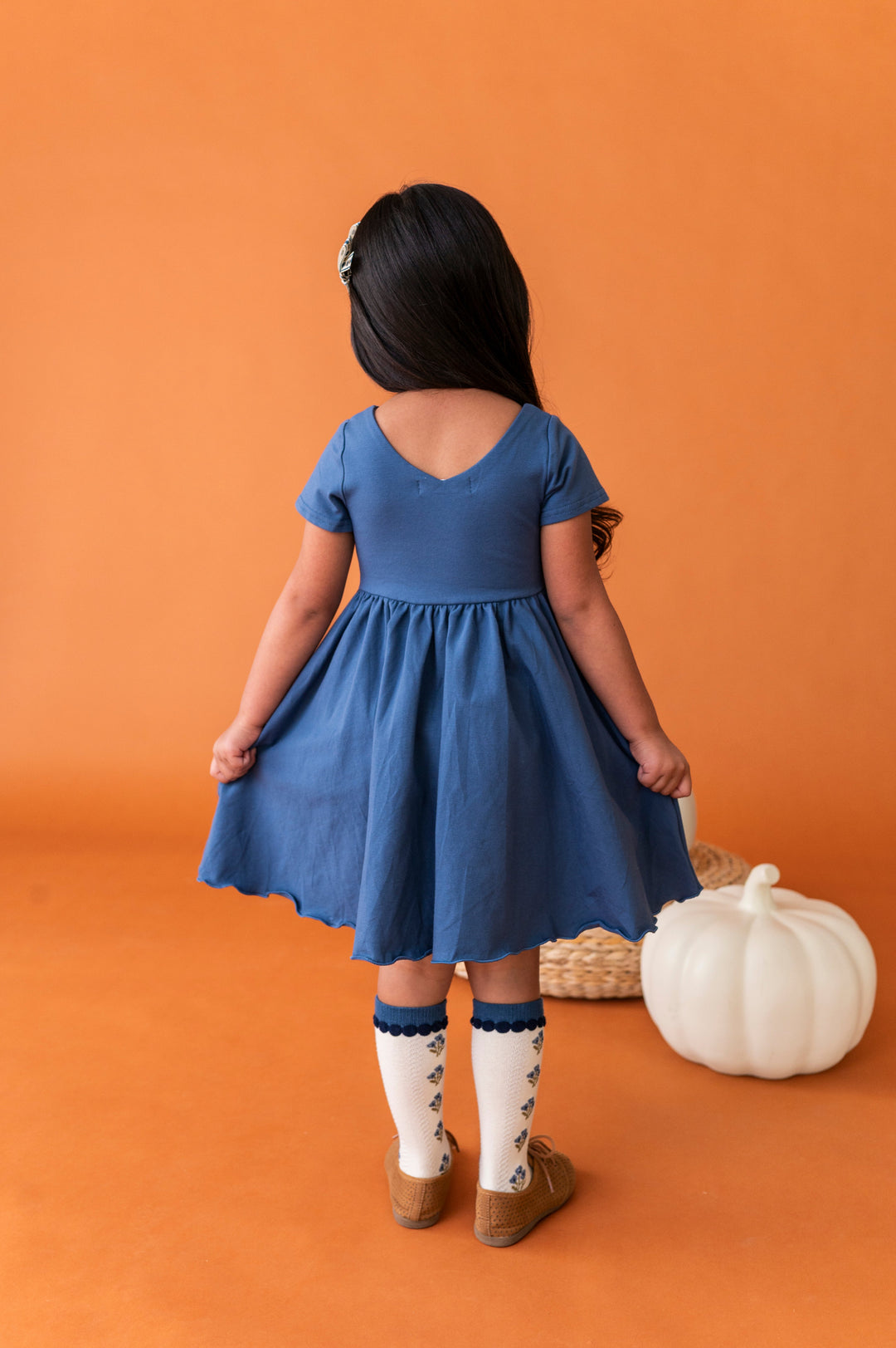 young girl facing away from camera next to white pumpkin to show v-neck design on back of blue cotton blend twirl dress