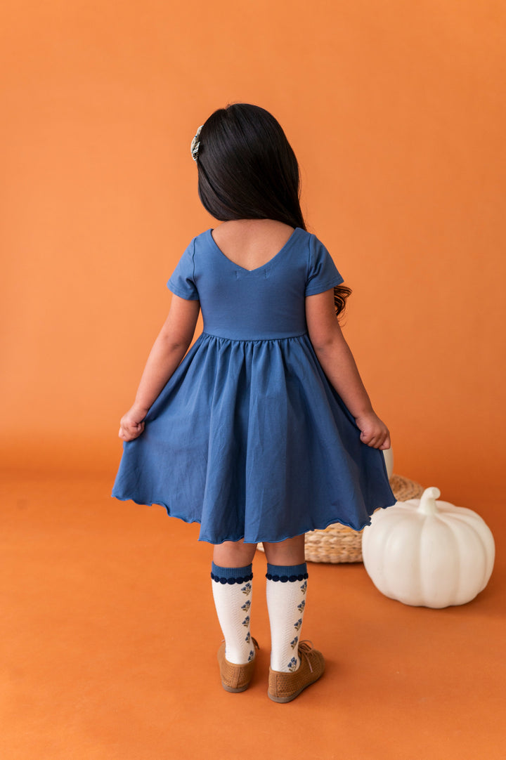 young girl facing away from camera next to white pumpkin to show v-neck design on back of blue cotton blend twirl dress