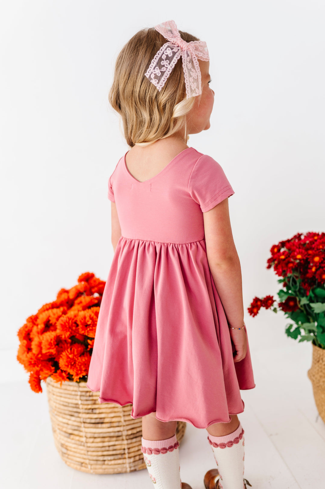 little girl showing v-neck design on back of rosy, mauve cotton blend twirl dress paired with light pink lace bow and matching floral knee socks