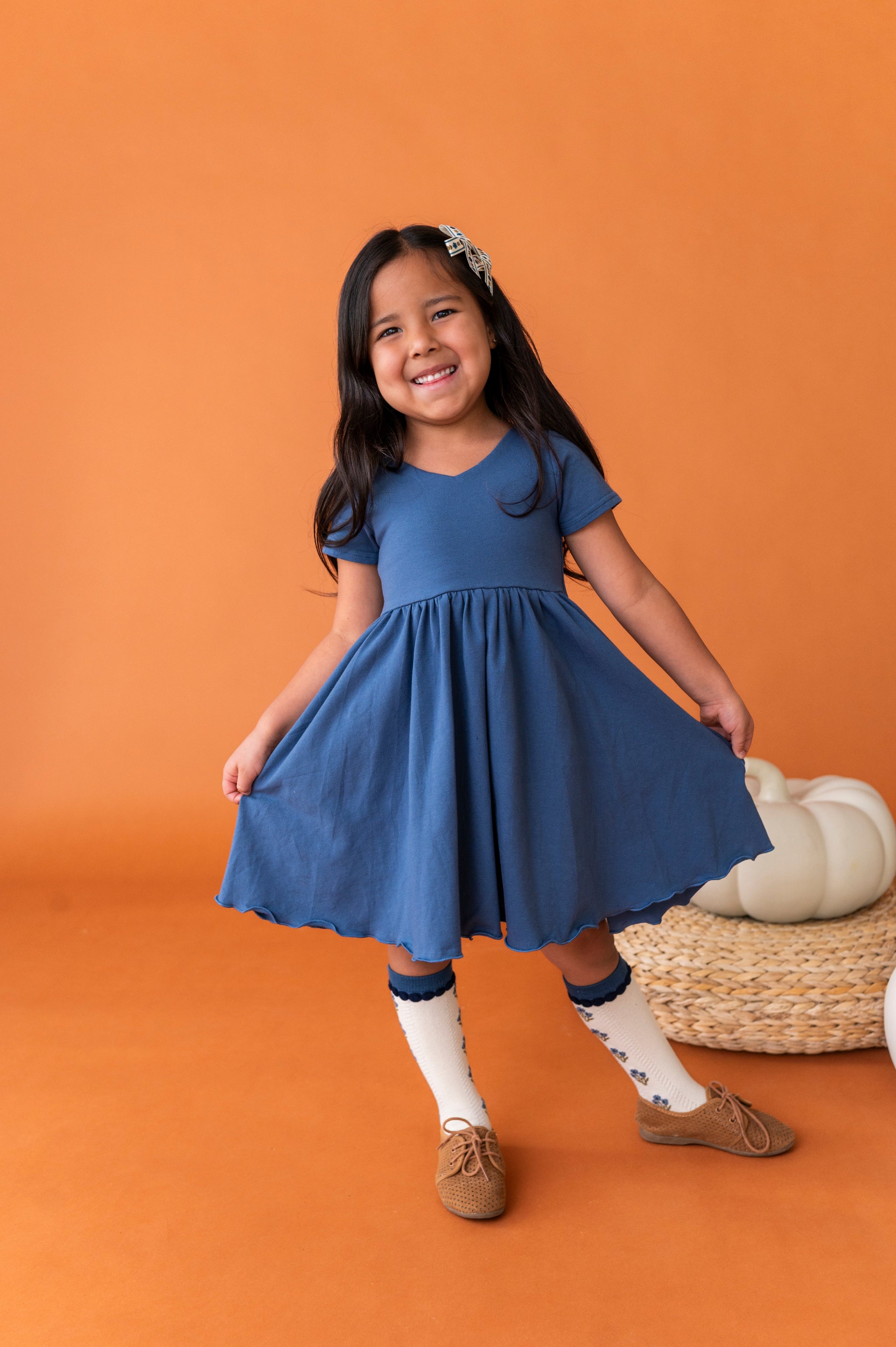 young girl near white pumpkin in solid fall blue v-neck twirl dress paired with matching blue floral knee socks and pigtail bows