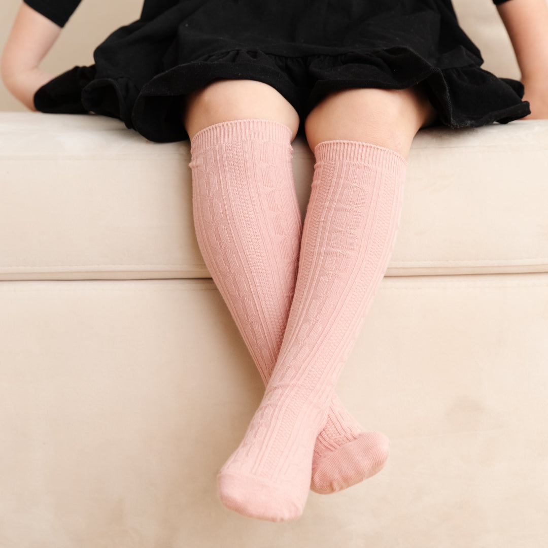 little girl sitting with crossed legs wearing blush pink cable knit knee high socks by little stocking co