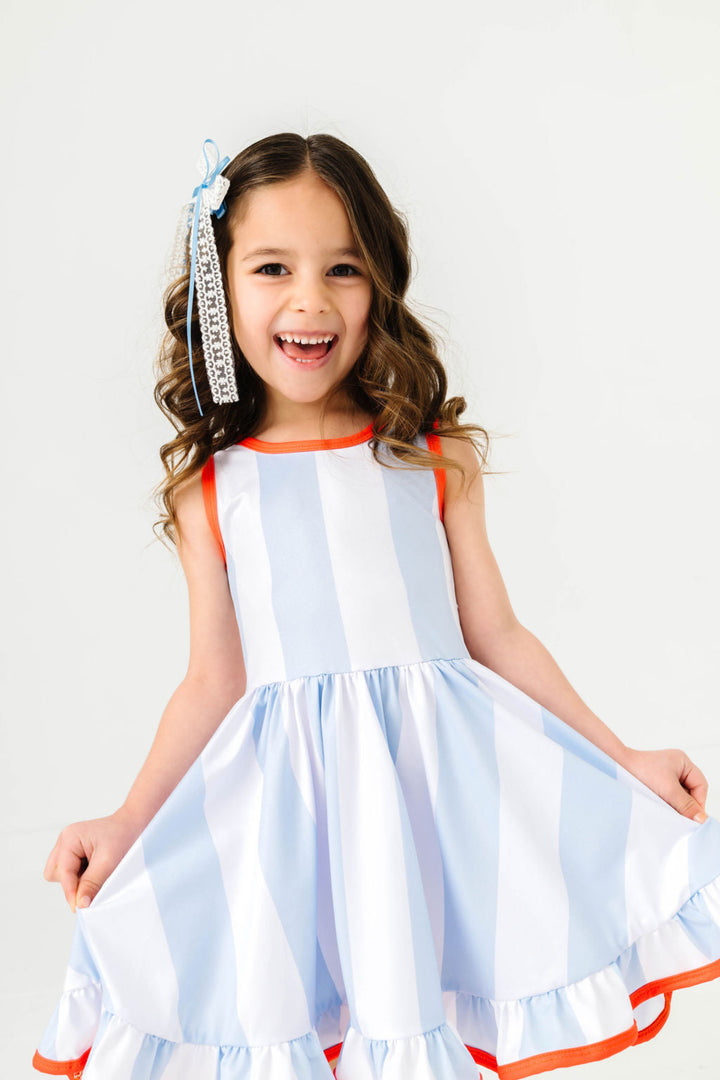 little girl posing in light blue and white wide vertical stripe sleeveless twirl dress with orange trim and white and blue lace bow