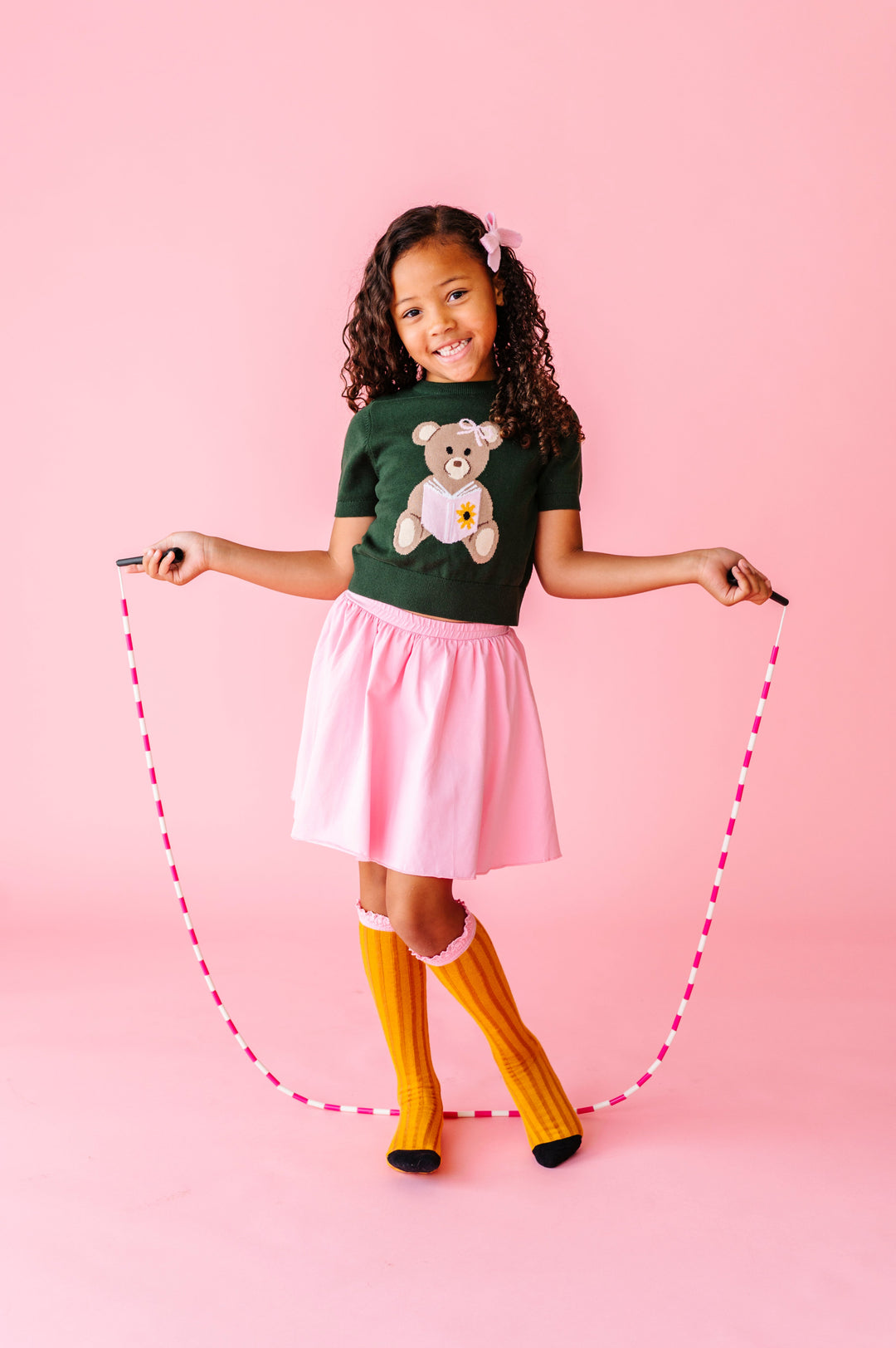 girl with jump rope wearing green school sweater with bear reading book paired with matching pink skort and pencul knee socks