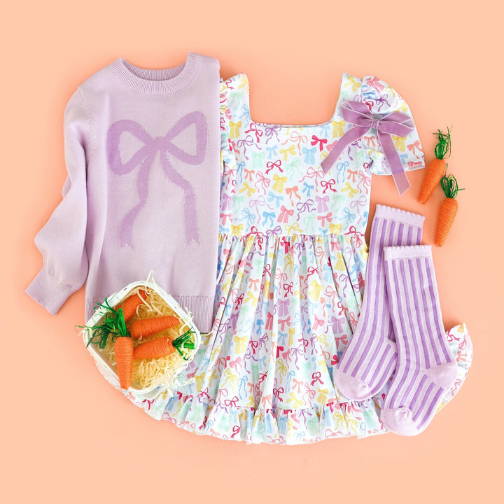 flatlay of girls white twirl dress with pastel colored bw print and matching lilac bow sweater and striped purple socks and lavender velvet bow