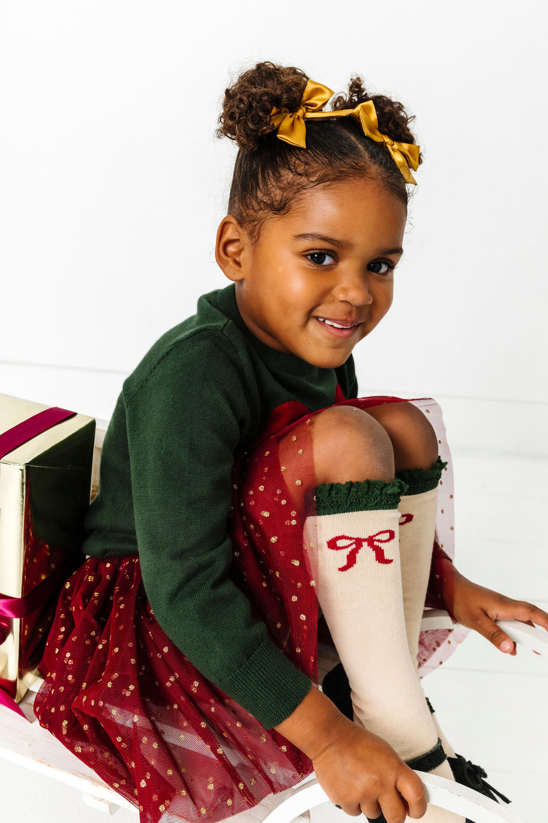 girl on sled wearing crimson and forest green Christmas outfit paired with gold satin pigtail bows and bow knee highs with green lace trim