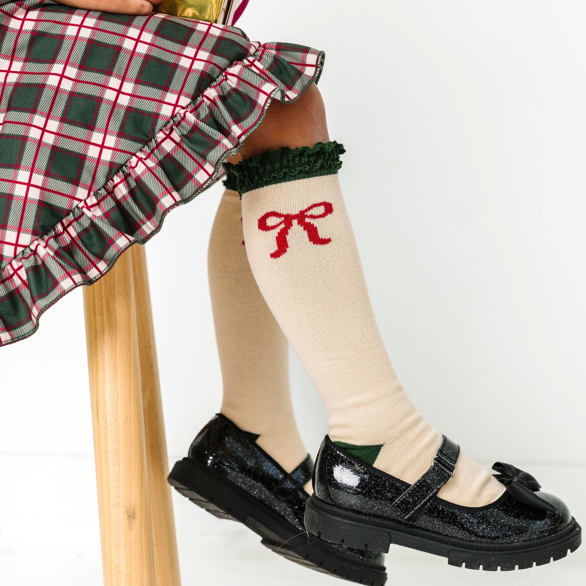 girl sitting with Christmas gift in cream knee high socks with green trim and red bow design paired with matching plaid dress