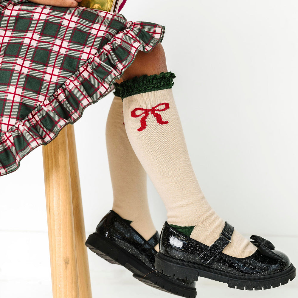 girl sitting with Christmas gift in cream knee high socks with green trim and red bow design paired with matching plaid dress