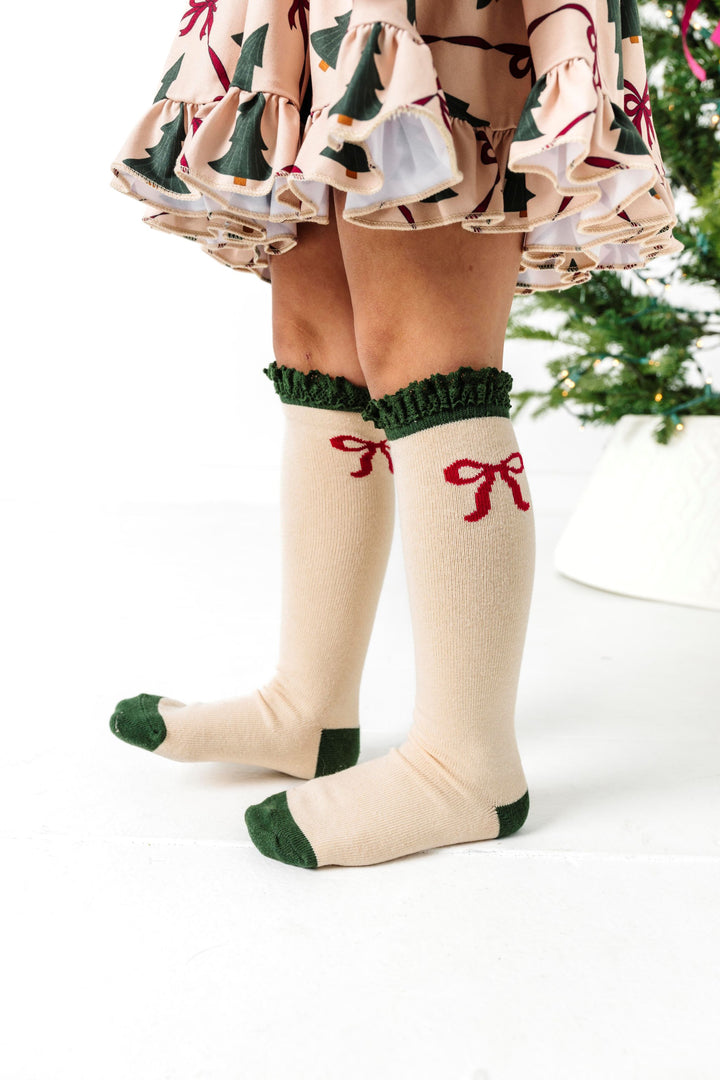 close up of cream knee high socks with forest grean heel, toe and lace trim and little crimson bow design paired with matching Christmas tree and bow dress