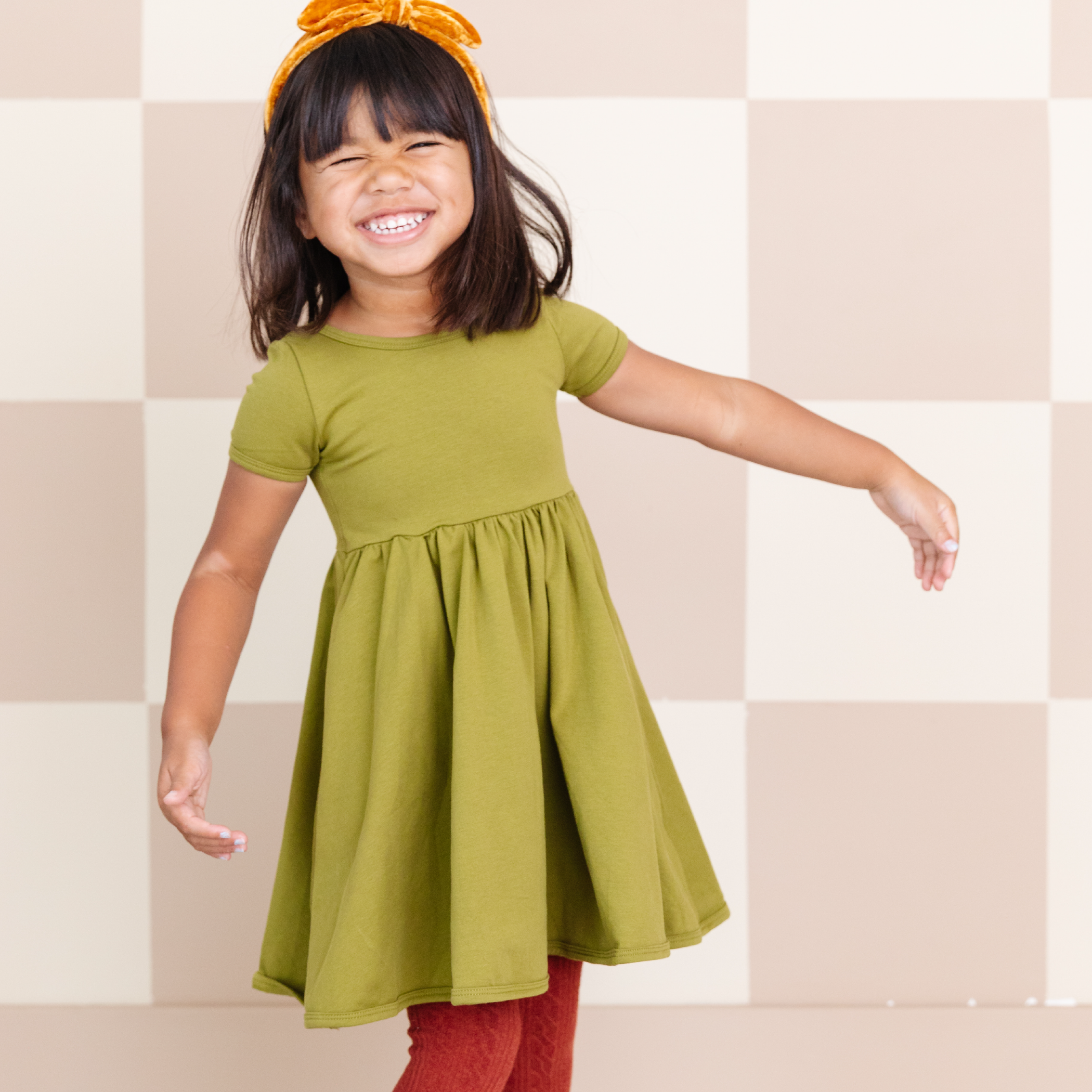 little girl wearing bright olive green cotton pocket dress and brick red tights and yellow velvet headband